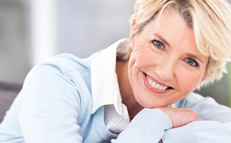 Eine lächelnde, reife Frau mit kurzen blonden Haaren, die in einem hellblauen Hemd gekleidet ist und entspannt auf einem Sofa lehnt. Ihr strahlendes Lächeln und ihr gesundes Aussehen könnten auf das Wohlbefinden hinweisen, das durch Hormonersatztherapie unterstützt wird.