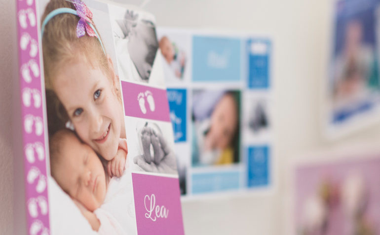 Verschwommenes Bild von einer Wand mit verschiedenen Fotos, die Kinder und Babys zeigen. Im Fokus steht ein Bild von einem Mädchen, das ein Baby im Arm hält, mit dem Namen "Lea" darunter. Die Darstellung könnte in einem Kontext stehen, der Wünsche nach Elternschaft oder die Freude über neue Familienmitglieder ausdrückt.