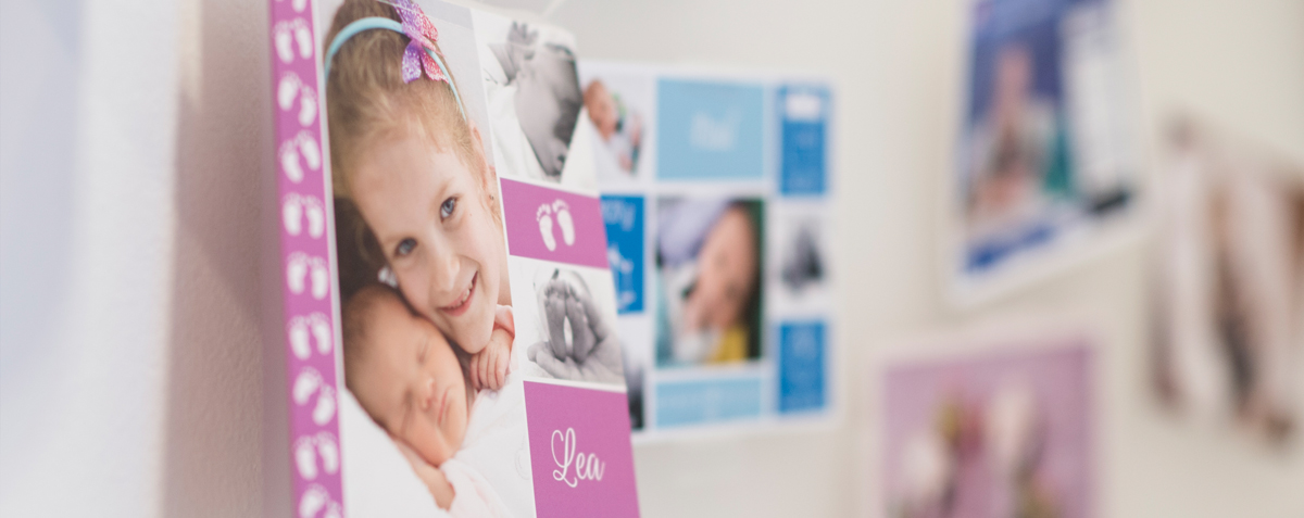 Verschwommenes Bild von einer Wand mit verschiedenen Fotos, die Kinder und Babys zeigen. Im Fokus steht ein Bild von einem Mädchen, das ein Baby im Arm hält, mit dem Namen "Lea" darunter. Die Darstellung könnte in einem Kontext stehen, der Wünsche nach Elternschaft oder die Freude über neue Familienmitglieder ausdrückt.
