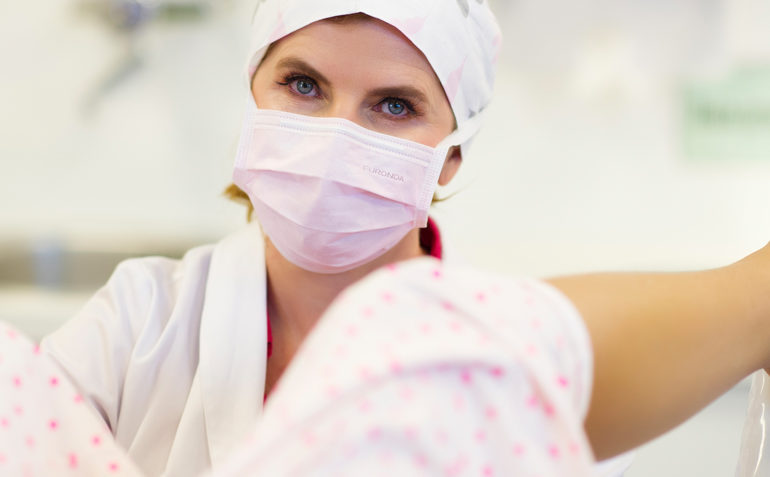 Eine Frauenärztin, Dr. Maria Riedhart-Huter, trägt eine Operationshaube und einen Mundschutz während einer gynäkologischen Untersuchung oder Behandlung. Im Vordergrund sind die Beine einer Patientin in medizinischen Beinhaltern zu sehen, die für einen Eingriff wie eine Schamlippenkorrektur vorbereitet sein könnten.