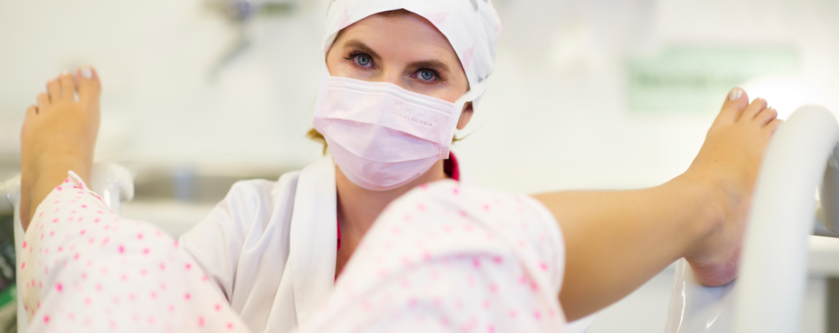 Eine Frauenärztin, Dr. Maria Riedhart-Huter, trägt eine Operationshaube und einen Mundschutz während einer gynäkologischen Untersuchung oder Behandlung. Im Vordergrund sind die Beine einer Patientin in medizinischen Beinhaltern zu sehen, die für einen Eingriff wie eine Schamlippenkorrektur vorbereitet sein könnten.