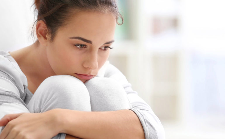 Eine Frau sitzt mit den Händen auf den Knien auf einem Bett und zeigt Anzeichen von Lichen sclerosus.